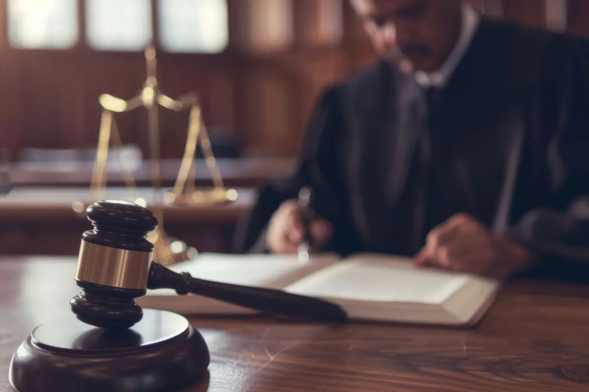 A judge is reading a book while holding a pen on his right hand. A justice scale and gavel with sound block is in front of the judge and on top of a wooden table.