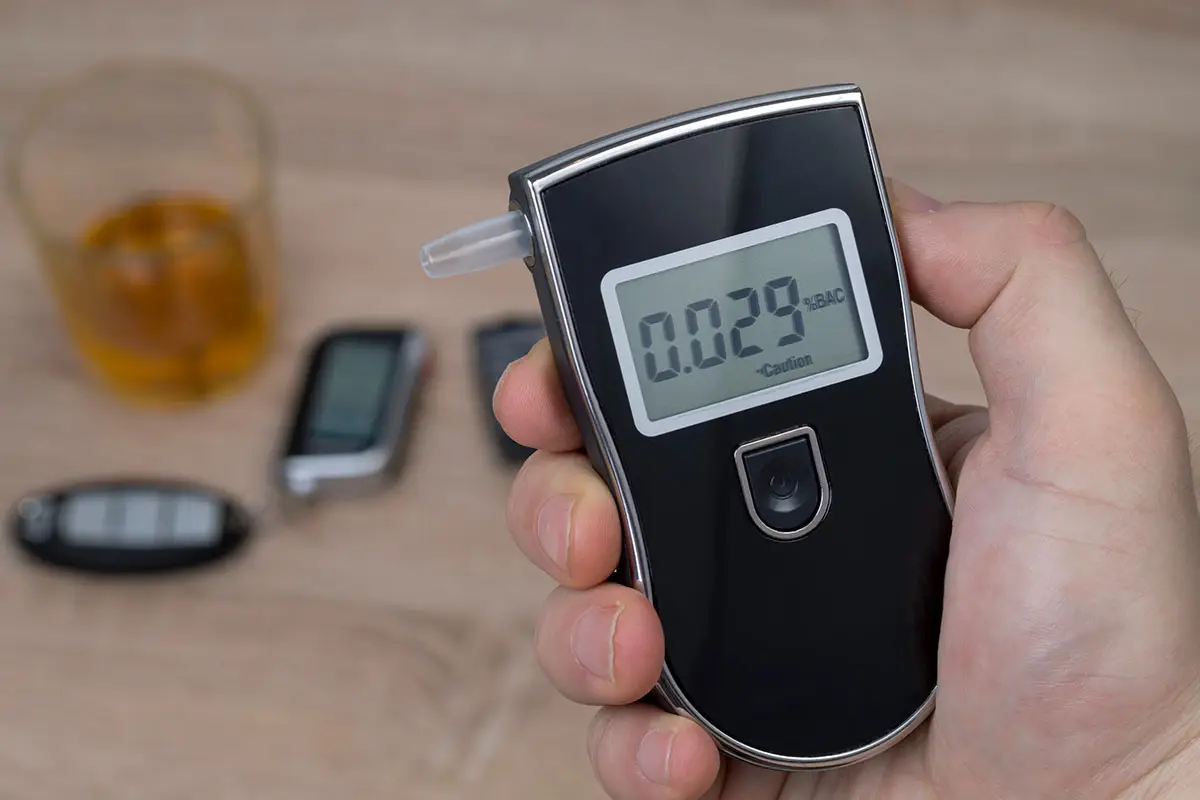 A man checks the results of a breathalyzer test. Car keys and a glass with liquor is in the background.