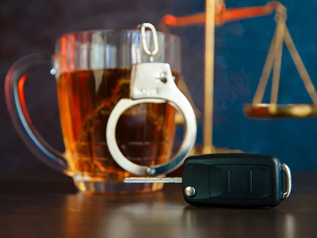 A glass with liquor, justice scale, handcuff, and car keys on top of a wooden table.