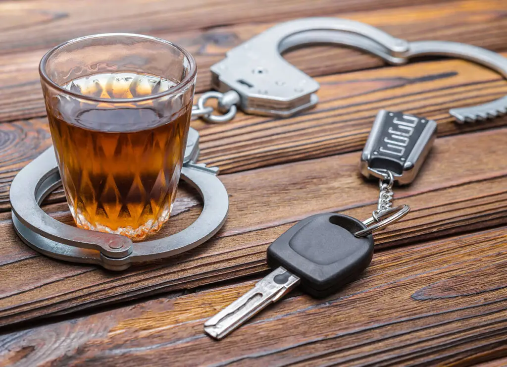 handcuffs, a glass with liquor, and car keys are on top of a wooden table.