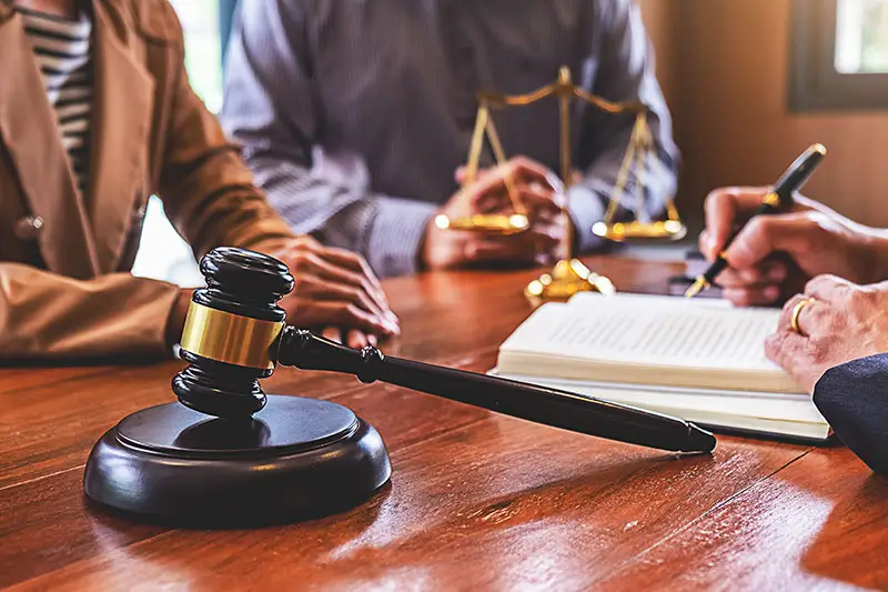 A lawyer speaks with his client as he writes on a notebook. A justice scale and gavel is on top of the wooden table.