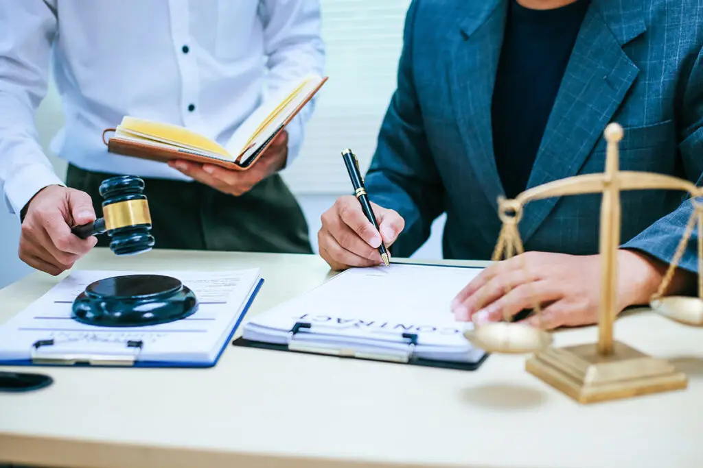 A client signing a document while the lawyer holds a book on his left hand and his right hand is about to pound the sound block of the gavel. A justice scale is adjacent the client.