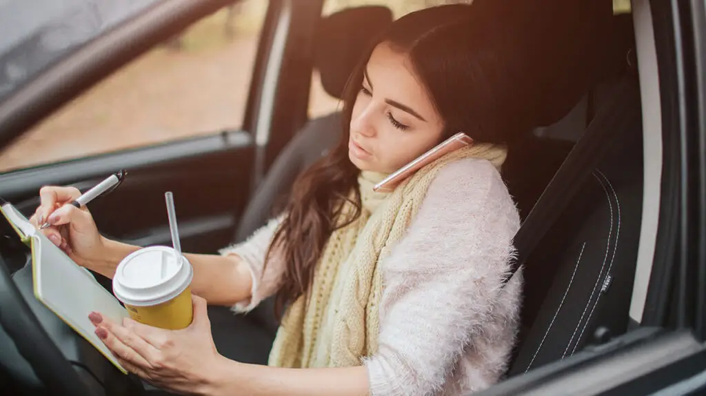 A woman is writing on her notebook with her right hand and holding a drink on her left hand while on the phone inside her car/